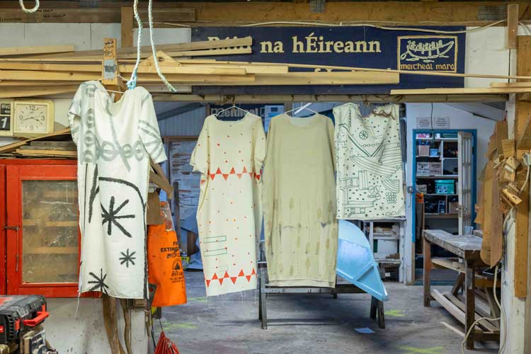 Garments from Ceyda Oskay’s clothing brand, cc by co, which she created for In From the Margins while an artist in residence at Cork Printmakers, hung in 2022 at Meitheal Mara Teoranta in Cork, the boatbuilding workshop that has employed former prisoners and others. The wheat depicted on the garment third from left is Red Stettin, which originated from the grain the Ottoman Empire sent to Ireland in 1847 during the Great Hunger. Photo courtesy John Beasley, Cork, 2022.