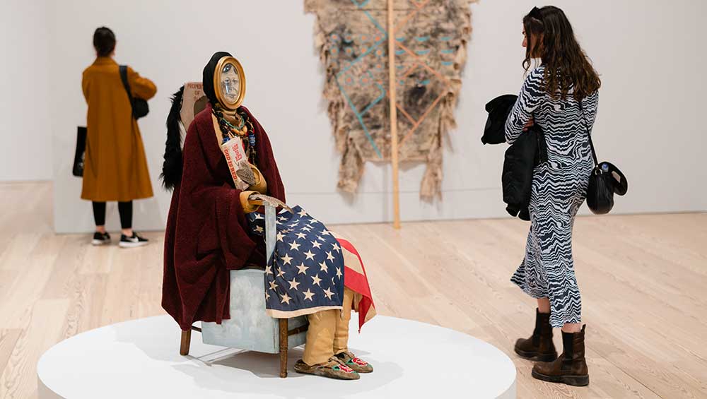 Jaune Quick-to-See Smith, Indian Madonna Enthroned, 1974. Burlap, fabric, polyester batting, dried corn, leather thongs, beaded leather bands, necklaces, book (God Is Red by Vine Deloria Jr.), pheasant wings, American flag, beaded hide moccasins, two framed ink and graphite pencil drawings, Masonite cradleboard, animal hide, sheepskin and fleece, bird feet, wood chair, and painted plywood, 52 × 34 × 20 in (132.1 × 86.4 × 50.8 cm). Collection of the artist; courtesy Garth Greenan Gallery, New York. Fabricated by Andy Ambrose. © Jaune Quick-to-See Smith. Photo: Neal Ambrose-Smith.