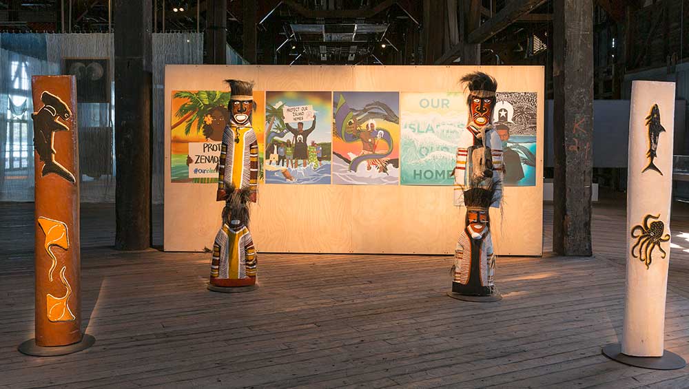 Yessie Mosby, Maluw Adhil Urngu Padanu Mamuy Moesik (Legends from the deep, sitting peacefully upon the waters), 2022. Courtesy the artist. Co-commissioned by the Biennale of Sydney and The Institute of Modern Art, Brisbane; and Torres Strait 8, Poster wall, 2022 (detail). Courtesy the artists with posters by Mooki Pen, Dylan Mooney, Guy Ritani, BlakSeed, Waniki Maluwapi, Jaelyn Biumaiwai. Photography by Mary Harm, Bindimu, Torn Parachute and Daniel Billy. Co-commissioned by the Biennale of Sydney and The Institute of Modern Art, Brisbane. Background: Clare Milledge, Imbás: a well at the bottom of the sea, 2022 (detail). Courtesy the artist & STATION. Commissioned by the Biennale of Sydney with generous support from the Australia Council for the Arts. Installation view, 23rd Biennale of Sydney, rīvus, 2022, Pier 2/3 Walsh Bay Arts Precinct. Photography: Document Photography.
